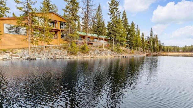 A lodge with a wooden exterior, a balcony, and a view of a calm lake surrounded by trees. - Lakefront Suite w/ Private Hot Tub | Fireplace (Naramata)