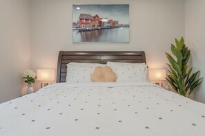 Un lit bien fait, recouvert d’une couvre-lit à motifs, agrémenté d’un coussin décoratif et orné d’un tableau représentant un paysage de bord de mer, accroché au mur.