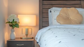 A neatly made bed with a large decorative pillow, a bedside lamp, and a vase with flowers on a nightstand. - Enjoy this Brand New Cozy 2BR Guest Suite in YYC! (Calgary)
