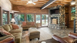 A spacious living room with a stone fireplace, leather sofas, and large windows offering a view of the outdoors. - Spacious Riverside Lodge on 370 acres near Tuscaloosa, 8 bedrooms - sleeps 15 (Gordo)