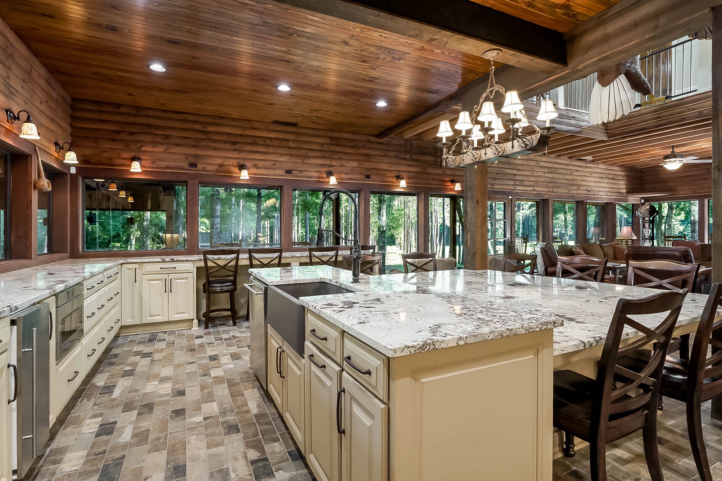 A spacious kitchen with a large island, wooden ceiling, and large windows offering a view of the outdoors.