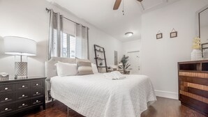 Ein Schlafzimmer mit einem Bett, einem Schminktisch, einer Lampe, einem Deckenventilator und einem Fenster mit Vorhängen.