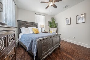 Quarto com cama de madeira, ventilador de teto, mesa de cabeceira, luminária e planta em vaso.
