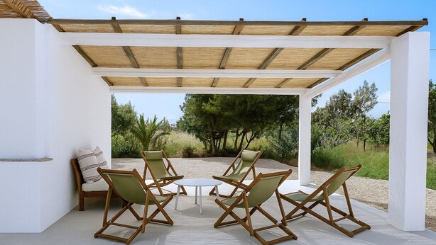 A covered outdoor seating area with four wooden chairs and a small table, surrounded by white walls and a view of trees and shrubs. - Mas Midó. Masia Restaurada en Delta del Ebro (Sant Jaume d'Enveja)
