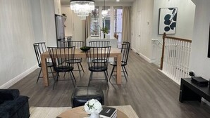Ett modernt vardagsrum med en matplats, en svart lädersoffa, ett träbord med en vas och böcker, och en trappa med ett räcke.