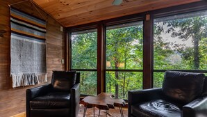 Une pièce chaleureuse aux murs en bois, avec une grande fenêtre donnant sur les arbres, deux fauteuils en cuir et une petite table en bois.