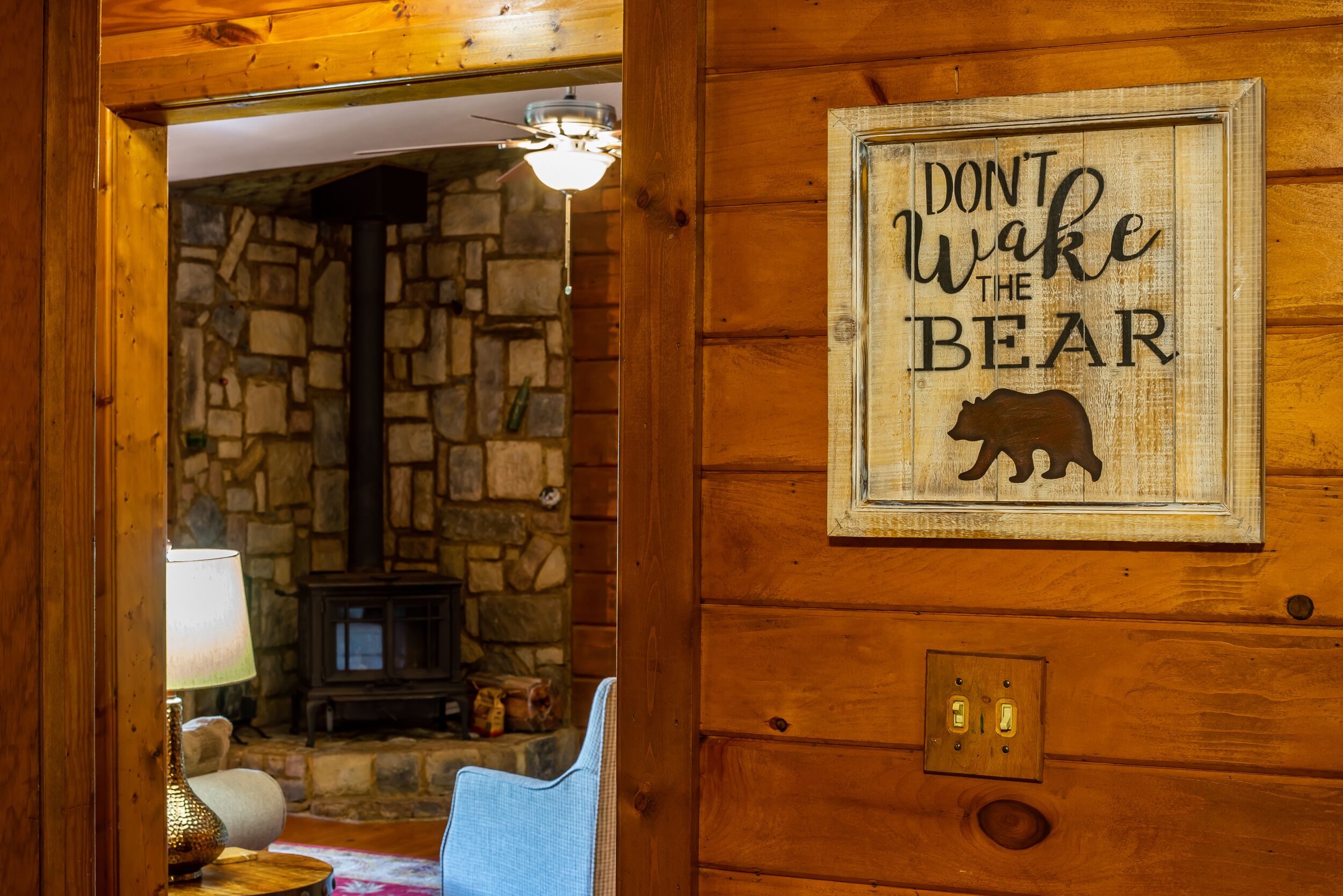 Et træinteriør med en stenvægge og pejs, et indrammet skilt med teksten "Don't Wake the Bear" og en blå stol.