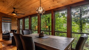 Une table à manger en bois, des chaises, un ventilateur de plafond et de grandes fenêtres donnant sur la forêt.