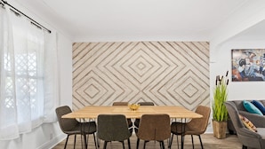 Un coin repas avec une table en bois et six chaises, un mur décoré d’un motif, un canapé et une plante en pot.