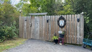 Une clôture en bois ornée d’un panneau décoratif en forme de lettre « W », un pot de fleurs et une brouette.