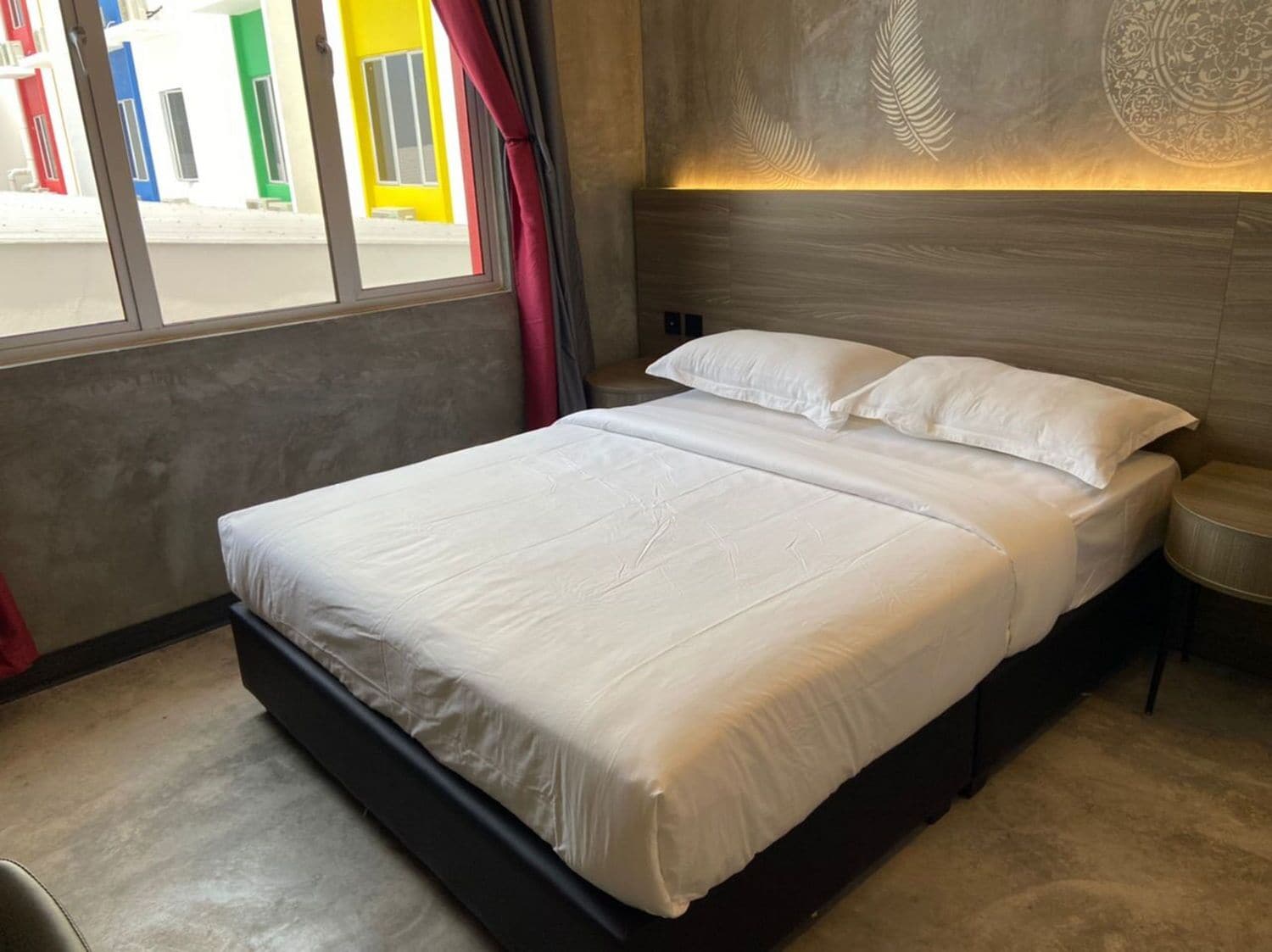 Cama doble con sábanas blancas en una habitación moderna con pared de concreto y un diseño decorativo con hojas.
