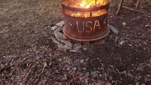 A man sitting by a fire pit with the word "USA" on it, surrounded by a yard with trees and a house in the background. - Shady Rest in Hot Springs MT. (Hot Springs)