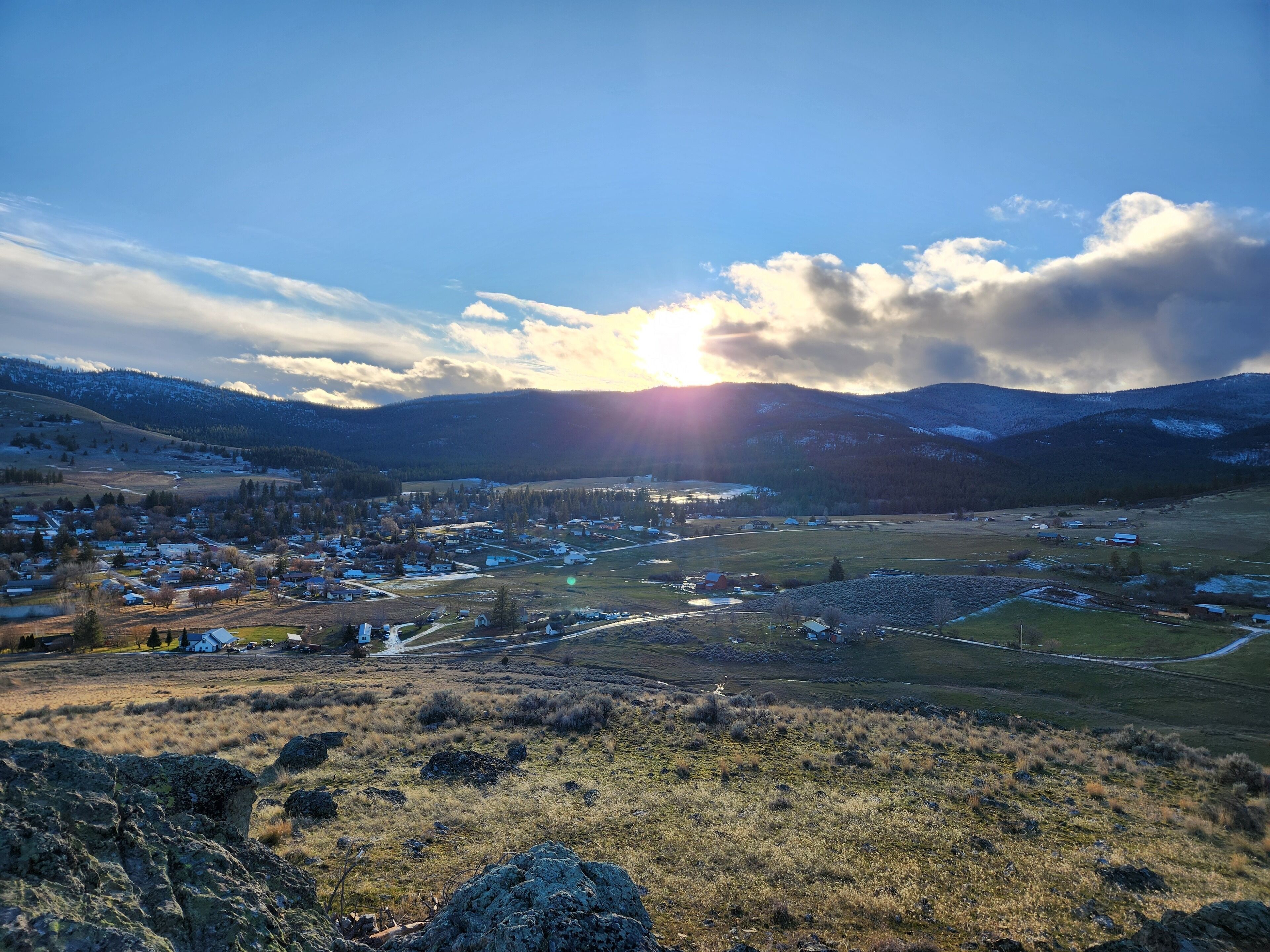 A panoramic view of a town with a river, surrounded by mountains and fields.