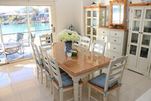 Un coin repas avec une table en bois et des chaises blanches, un vase de fleurs et une vue sur une piscine et des bâtiments.