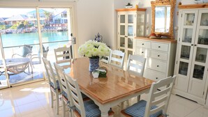 A dining area with a wooden table and white chairs, a vase of white flowers, and a view of a pool and buildings outside.
