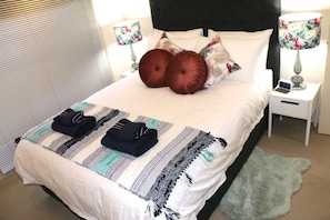 Un lit bien fait, recouvert d’une couverture à rayures, avec deux oreillers et une serviette pliée posée sur le dessus. On y trouve deux lampes de chevet avec des abat-jour à motifs floraux, une petite table de chevet avec une horloge, et un tapis vert sur le sol.