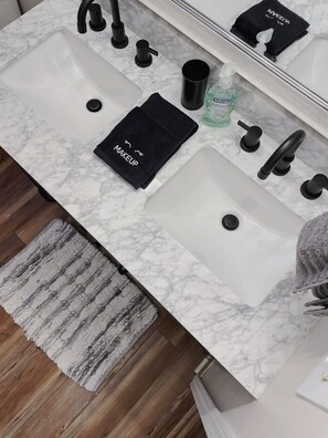 Ein Badezimmer mit einem weißen Marmorwaschbecken, schwarzen Wasserhähnen, einem schwarzen Handtuch mit der Aufschrift 'MAKEUP', einem grünen Seifenspender und einem Holzboden mit grauem Teppich.