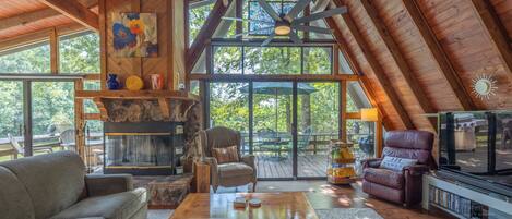 A spacious loft with a high ceiling, wooden beams, a fireplace, comfortable seating, and large windows offering a view of the outdoors.