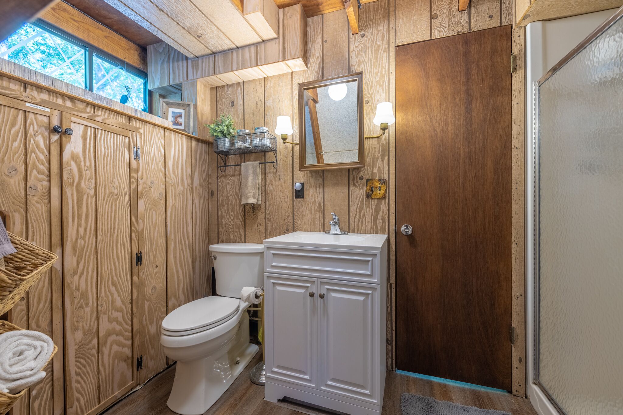Une salle de bain aux murs lambrissés, avec des toilettes, un lavabo blanc et un miroir au-dessus du lavabo.