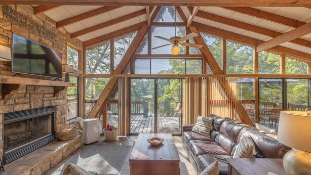 A spacious living room with a stone fireplace, leather sofa, and large windows offering a view of the outdoors. - The Firefly Inn by Innsbrook Vacations! (Innsbrook)