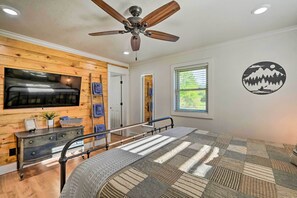 A bedroom with a bed, a ceiling fan, a TV mounted on a wooden wall, a window, and a wall decal. - Broken Bow Retreat w/ Large Yard + Fire Pit! (Broken Bow)