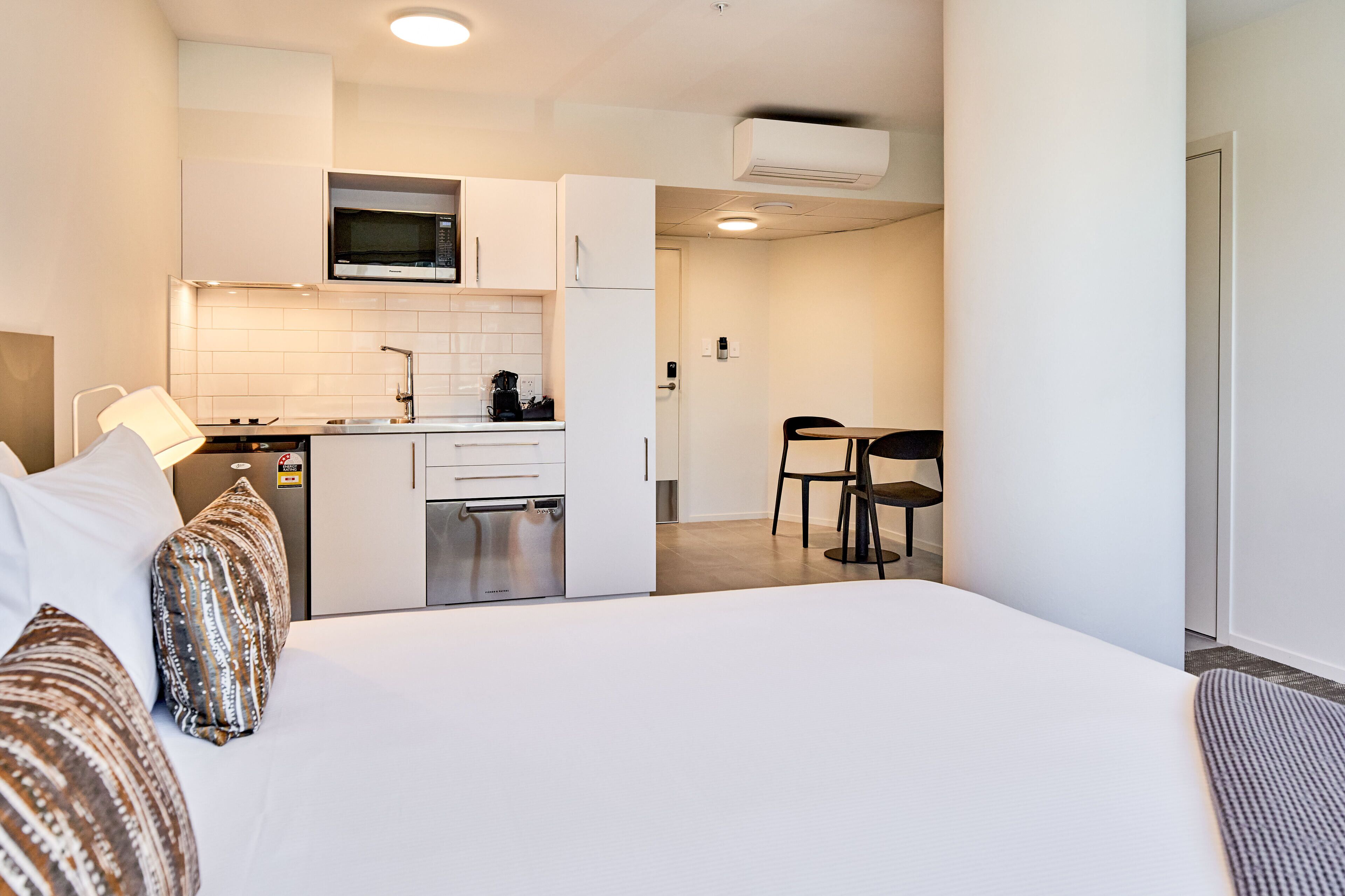 A modern hotel room with a large bed, a kitchenette, and a dining area.