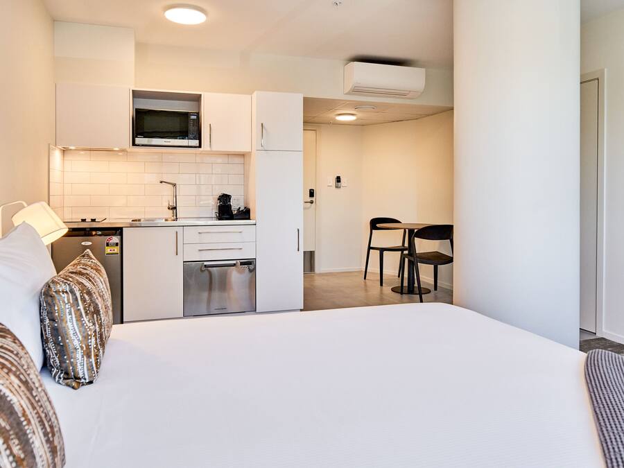 A modern hotel room with a large bed, a kitchenette, and a dining area.