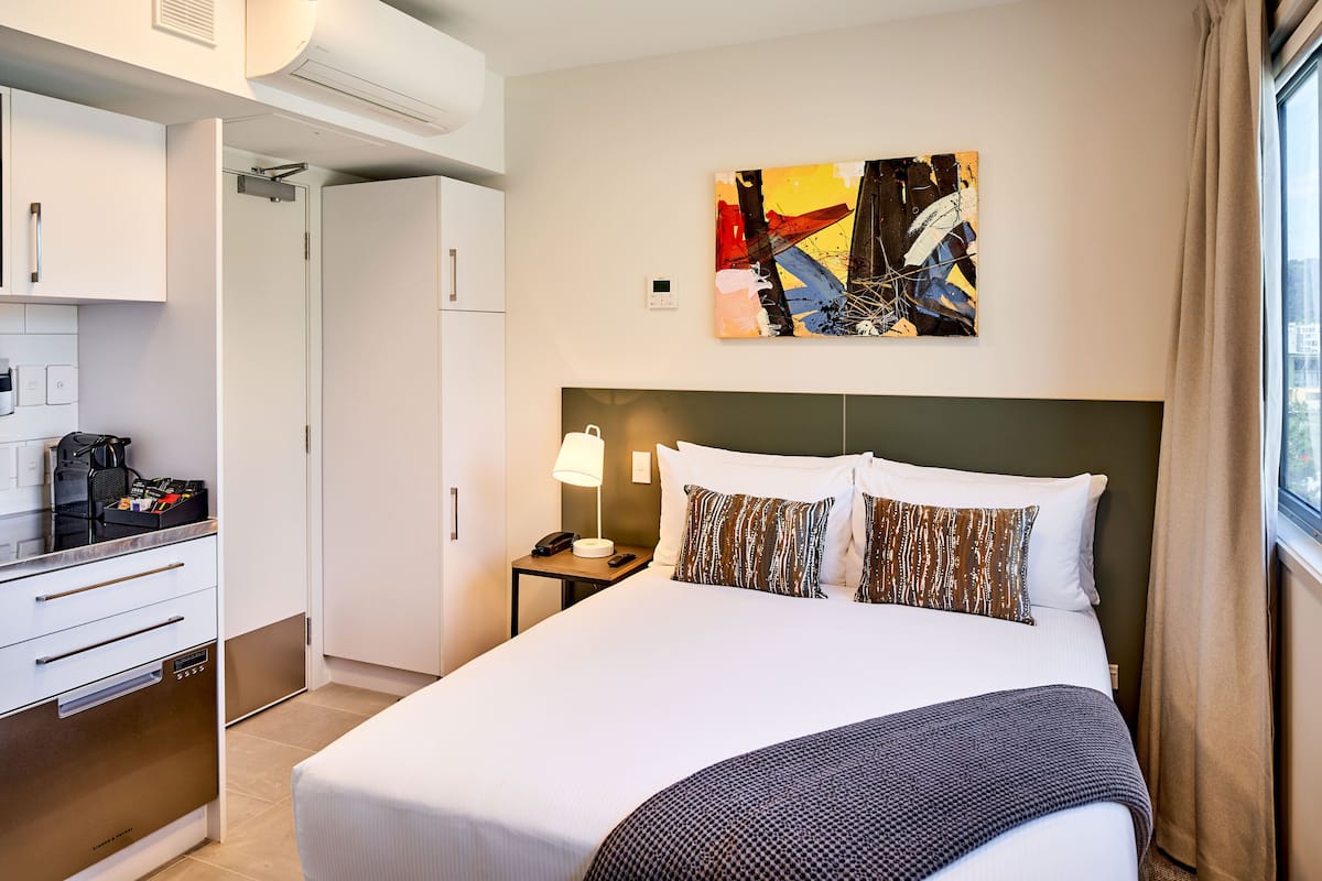 Une chambre d’hôtel avec un lit, une table de chevet, une lampe, un tableau et une kitchenette.