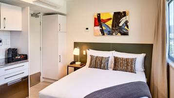 Quarto de hotel com cama, mesa de cabeceira, luminária, quadro e cozinha compacta.