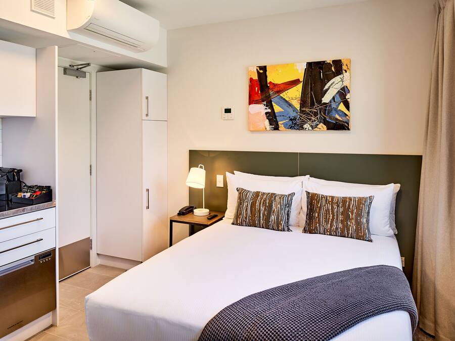 Quarto de hotel com cama, mesa de cabeceira, luminária, quadro e cozinha compacta.