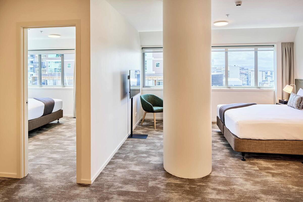 Une chambre d’hôtel avec un grand lit, un fauteuil vert, une télévision et une vue sur la ville grâce à de grandes fenêtres.