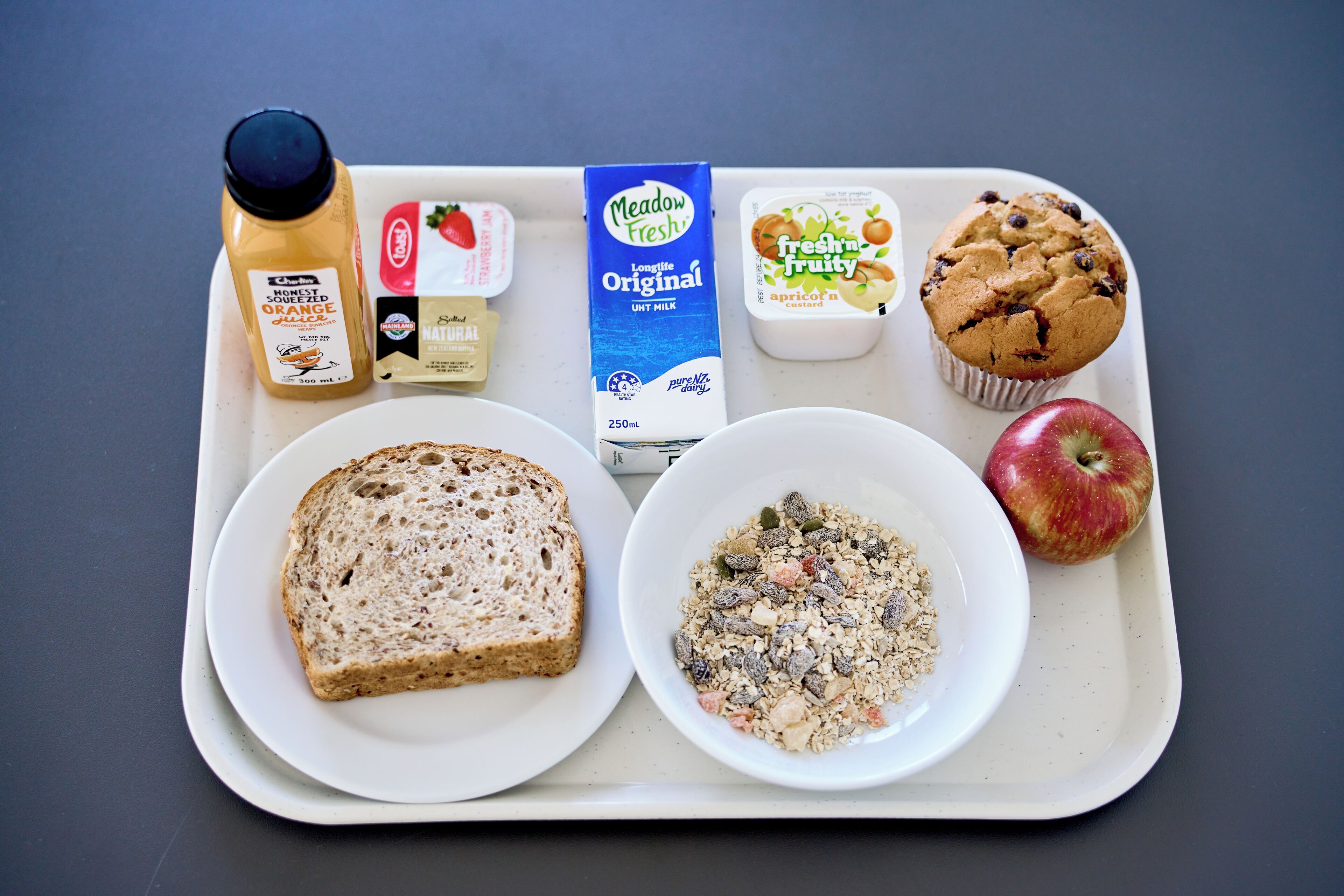 Una bandeja con una rebanada de pan integral, un tazón de granola, un muffin, una botella de jugo de naranja, un cartón de leche, un envase de yogur y una manzana.
