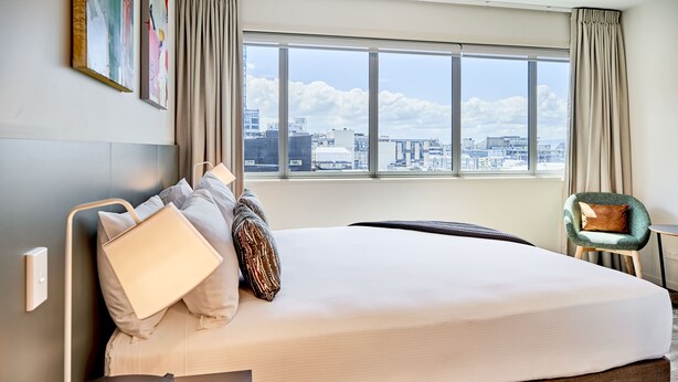A hotel room with a large bed, a desk, a chair, and a view of the cityscape through the window.