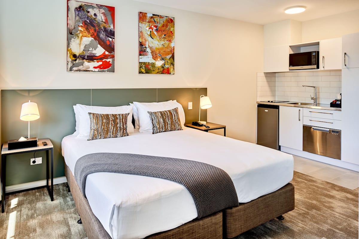 Une chambre d’hôtel dotée d’un grand lit, d’une table de chevet avec une lampe, d’un coin cuisine moderne et d’œuvres d’art abstraites accrochées au mur.