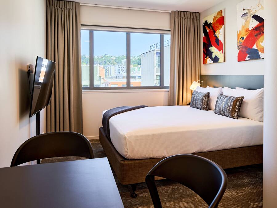 Habitación de hotel con cama, escritorio, silla, televisión y ventana con cortinas.
