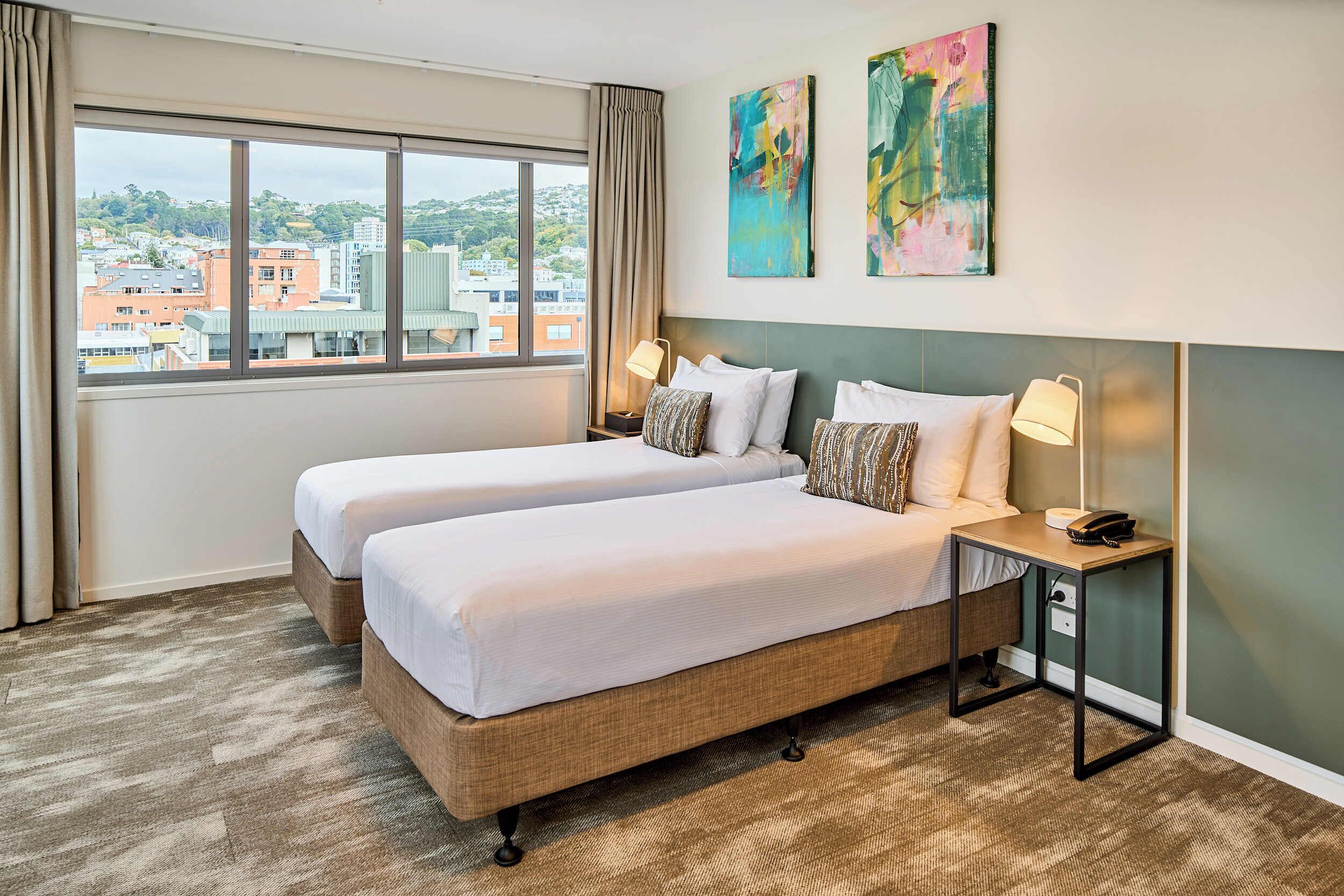 Habitación de hotel con dos camas, una mesita de noche con lámpara y vista a la ciudad a través de amplios ventanales.