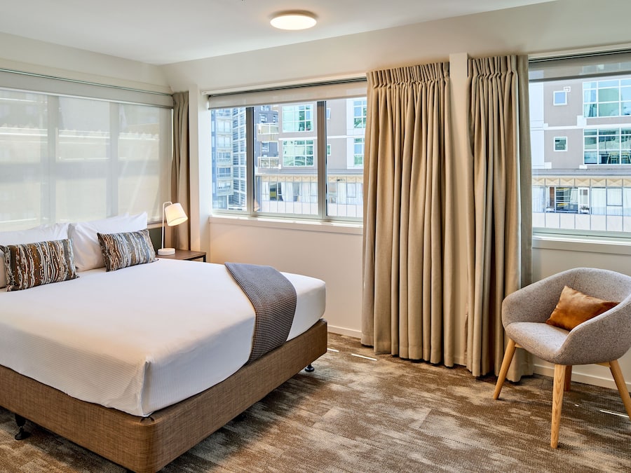 A hotel room with a large bed, a chair, and a view of buildings through the windows.