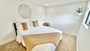 Un dormitorio con cama, almohadas, mesita de noche, lámpara, una obra de arte en la pared y una planta colgante.