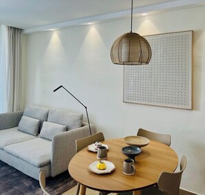 A modern living room with a sofa, a round dining table with chairs, a hanging light fixture, and a wall-mounted artwork.