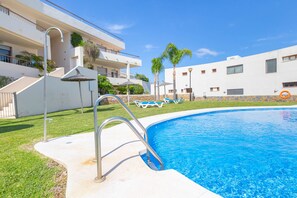 A swimming pool with a metal ladder, lounge chairs, and a building with balconies and greenery in the background.