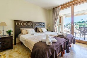 A hotel room with two beds, a nightstand, a lamp, a potted plant, and a view of the outdoors.