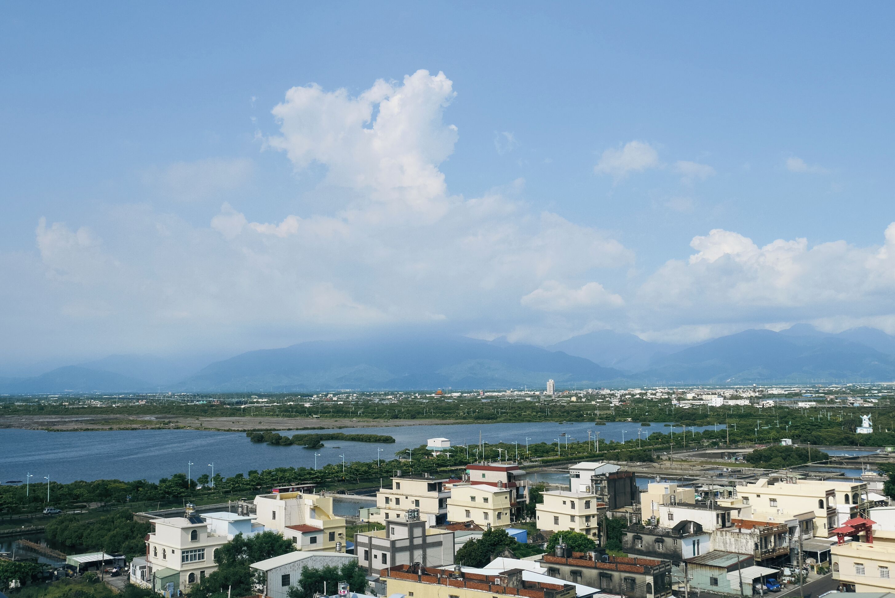 A coastal cityscape with buildings along the waterfront, a clear sky, and mountains in the distance.
