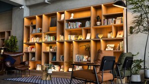 A modern library with multiple wooden bookshelves filled with books and decorative items. There are leather chairs and a small table with a patterned rug.