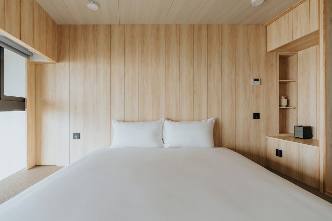 A bedroom with a wooden panel wall, a bed with white bedding, a nightstand, and a built-in shelf with a small appliance.