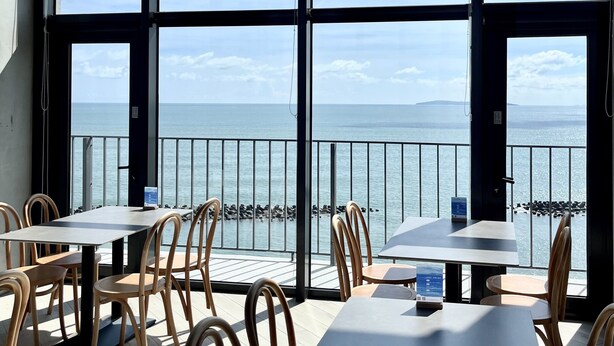 A dining area with tables and chairs, large windows with a view of the sea, and a balcony railing.