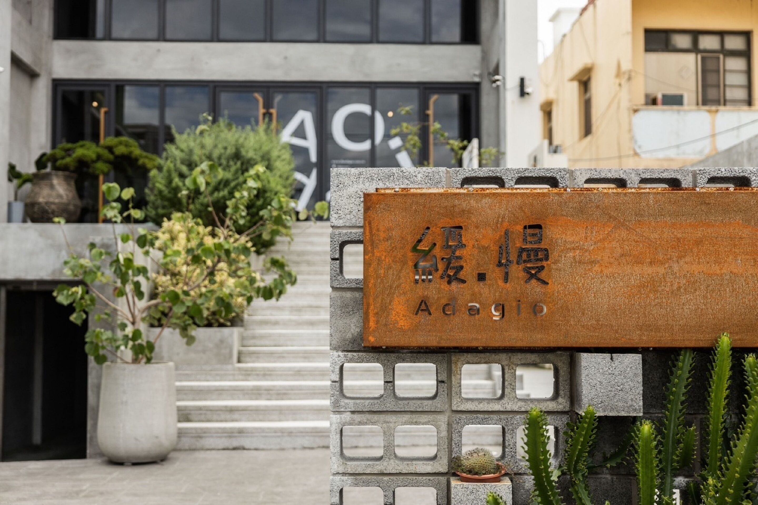 A building entrance with a sign in a foreign language, steps leading up to the entrance, and potted plants around the area.