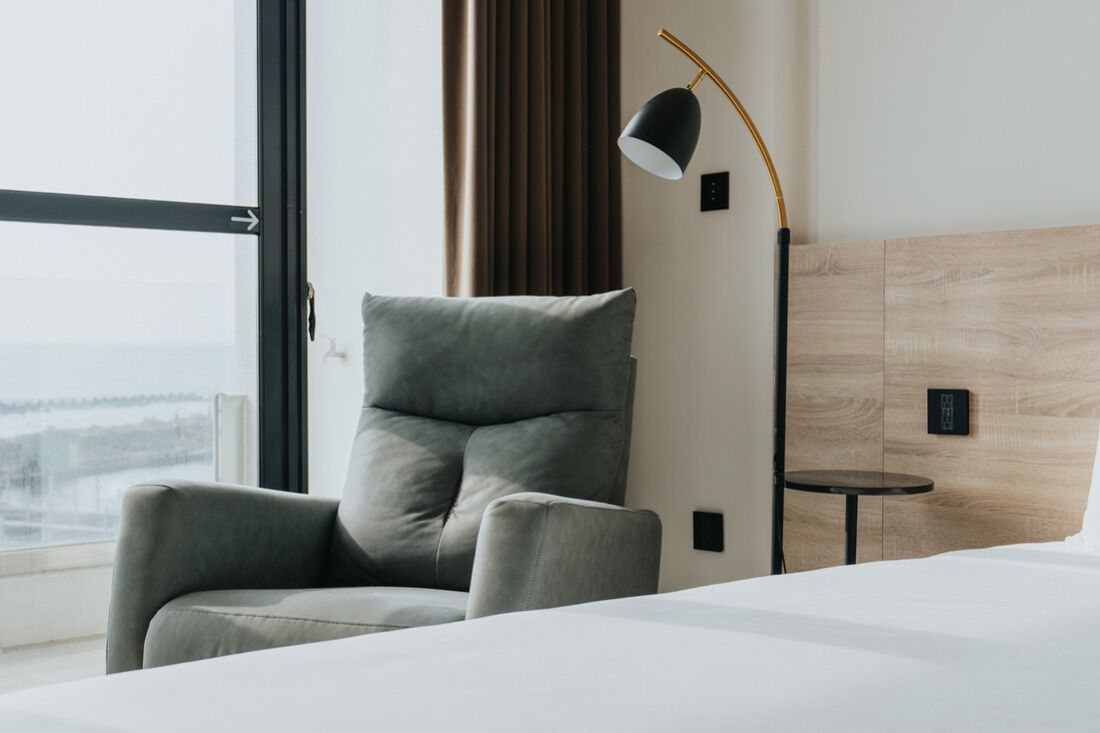 A modern hotel room with a large bed, a grey armchair, a small round table, and a floor lamp.