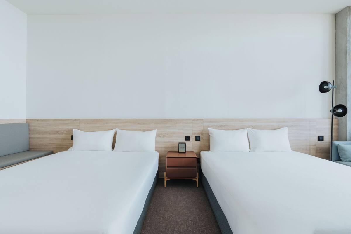 A hotel room with two beds, a nightstand with a clock, and a wall-mounted lamp.