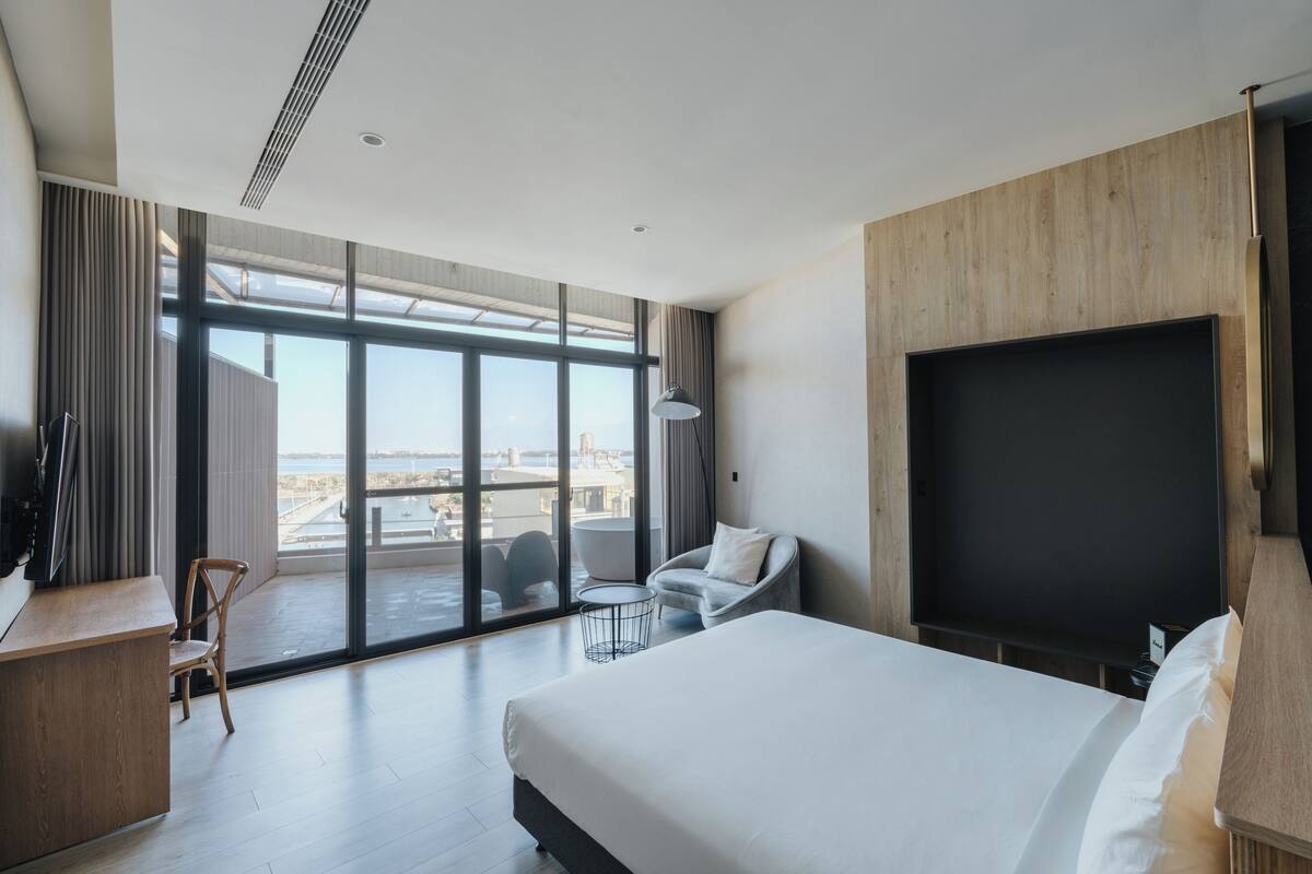 A modern hotel room with a large bed, a desk, a chair, a TV, and a balcony with a view of the city.