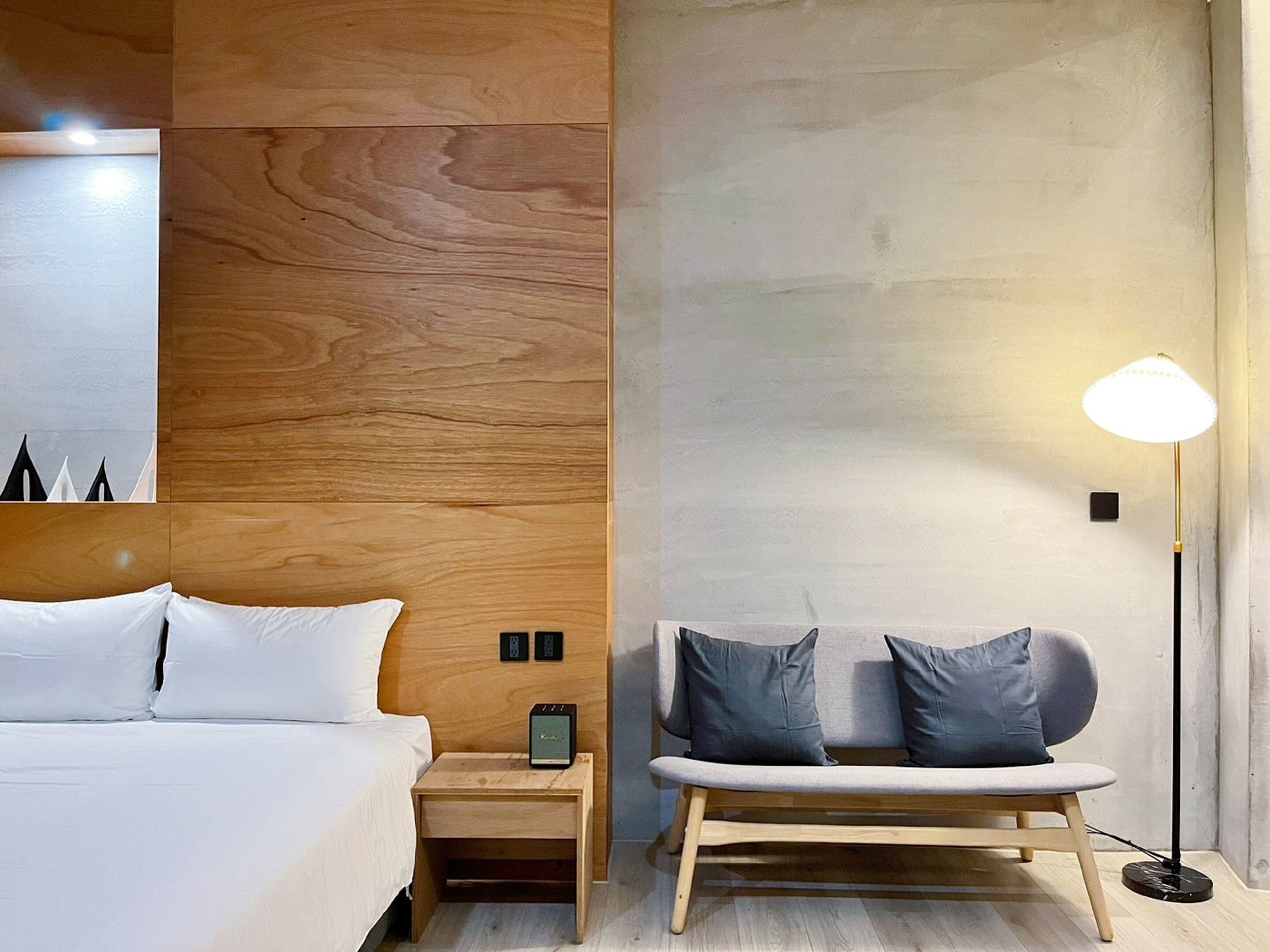 A modern hotel room with a wooden headboard, a bed with white linens, a bedside table with a lamp, and a bench with cushions.